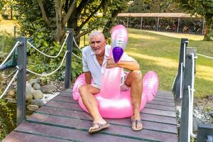retrato do sorridente bonito meio envelhecido homem sentado em a inflável flamingo brinquedo foto