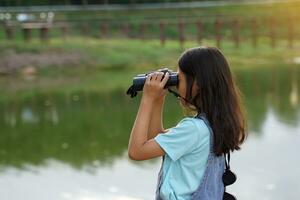 ásia menina usa binóculos para observar a natureza, pássaros, e animais do a nacional parque enquanto acampamento com dela família. com a conceito do Aprendendo lado de fora a Sala de aula natureza estudar, exploração. foto