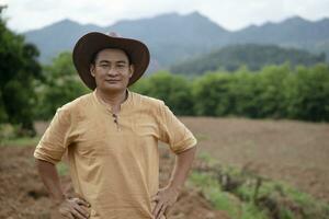 bonito ásia homem agricultor desgasta chapéu, Castanho camisa, colocar mãos em cintura, sente confiante, carrinhos às agricultura terra antes crescendo plantações. conceito, agricultura ocupação. tailandês agricultor estilo de vida. foto