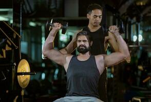 caucasiano homem é usando haltere dentro peso Treinamento para braço músculo dentro Academia com Apoio, suporte a partir de treinador para evita prejuízo Sombrio fundo para exercício e exercite-se conceito foto