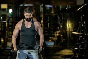 caucasiano barba muscular esporte homem é prática peso Treinamento em Duplo halteres para bíceps e tríceps músculo dentro Academia com Sombrio fundo para exercício e exercite-se foto