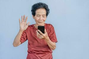 ásia Senior mulher sorrisos e vídeo ligar isolado dentro cinzento fundo. Forte idosos avó sentindo-me feliz usando Móvel célula telefone para comunicar com família e apreciar aposentadoria vida dentro casa. foto