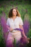 jovem mulher carrinhos dentro branco camisa dentro campo do roxa e Rosa tremoços. lindo jovem mulher com encaracolado cabelo e chapéu ao ar livre em uma Prado, tremoços florescer. pôr do sol ou nascer do sol, brilhante tarde luz foto