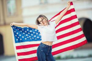 feliz jovem americano escola menina segurando e acenando dentro a cidade com EUA bandeira foto