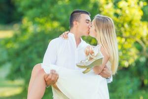 jovem homem carregando dele namorada dentro a braços enquanto se beijando dentro natureza em uma ensolarado dia.. ambos vestido dentro branco, ela detém dela sapatos dentro dela mão foto