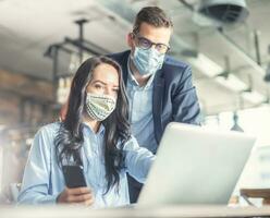 Boa olhando fêmea e masculino colegas de trabalho Veja às a computador tela junto, vestindo face máscaras foto