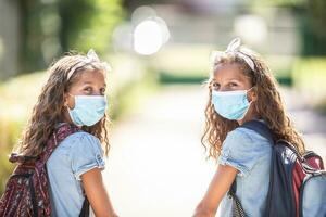 gêmeo irmãs com face mascarar costas às escola durante covid-19 quarentena foto