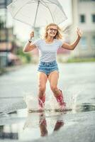 alegre menina pulando com branco guarda-chuva dentro pontilhado vermelho galochas. quente verão dia depois de a chuva mulher pulando e espirrando dentro poça foto