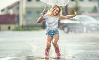 alegre menina pulando com branco guarda-chuva dentro pontilhado vermelho galochas. quente verão dia depois de a chuva mulher pulando e espirrando dentro poça foto