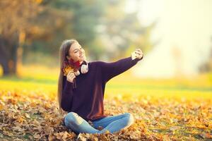 retrato do feliz jovem menina com fones de ouvido e Smartphone dentro outono parque ouvindo música ou faz selfie foto