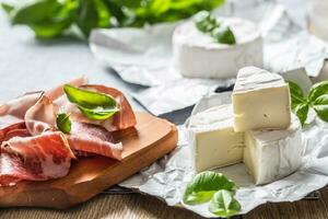 queijo Camembert ou brie queijo e carne de porco presunto prosciutto com manjericão folhas em mesa foto