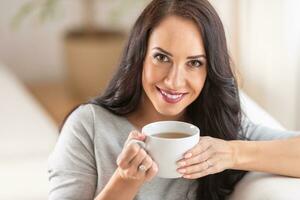 Sombrio cabelos mulher desfrutando uma adorável copo do chá ou café enquanto sentado em uma sofá. foto