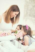 feliz mãe dia conceito. mãe com dois fofos jovem filhas gêmeos em a cama dentro a quarto e uma ramalhete do flores tulipas foto