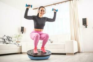 em forma lindo mulher dentro uma Treinamento equipamento e fones de ouvido agachamentos às casa em uma Saldo bola segurando dumbells para cima foto