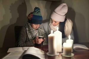 preocupado mãe confortos dela congelando filho segurando chá e estudando debaixo Luz de velas dentro a guerra zona, ambos vestindo inverno roupas às casa foto