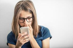 fofa jovem menina dentro azul camisa lê mensagem ou vídeo para Móvel telefone foto