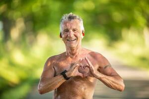 velho mas em forma homem com cinzento cabelo mostra uma coração gesto com dele dedos Como uma placa do dele saudável cardiovascular sistema foto