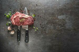 cru carne bife com garfo e faca sal Pimenta e ervas em Sombrio concreto fundo foto