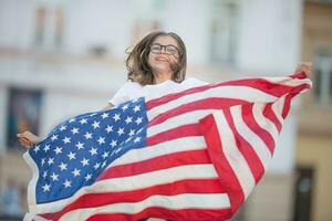 feliz jovem americano escola menina segurando e acenando dentro a cidade com EUA bandeira foto