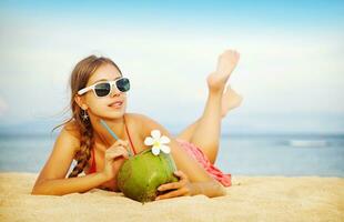 uma mulher dentro oculos de sol e uma vermelho vestir é deitado em a de praia com uma coco foto