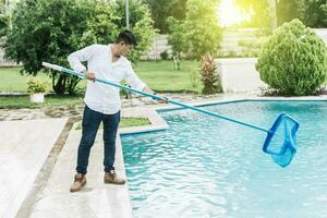 uma homem limpeza piscina com folha skimmer. homem limpeza a piscina com a escumadeira, pessoa com skimmer limpeza piscina, mãos segurando uma skimmer com azul piscina dentro a fundo foto