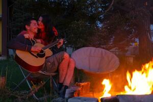 uma homem tocam a guitarra, uma mulher escuta e canta junto. uma casal dentro amor é sentado de a ao ar livre fogueira dentro a pátio do a casa em acampamento cadeiras, uma romântico tarde foto