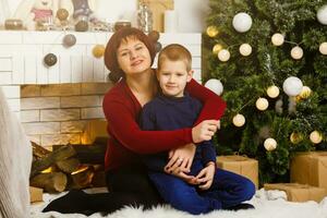 dentro a vivo quarto com uma lareira e uma Natal árvore, mãe e dela filho estão embalagem uma presente para Natal foto