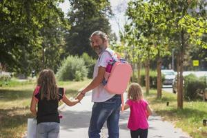 pai está levando as filhas para a escola, usando a mochila das crianças nas costas