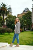 lindo jovem mulher vestindo Jaqueta e jeans caminhando em a calçada. Palma árvores e histórico construção fachada em fundo. cidade lazer. despreocupado pessoa em repouso experimentando calma foto