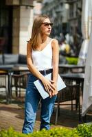 Maravilhoso jovem empresária em pé perto rua cidade cafeteria. menina vestindo branco tanque topo e jeans segurando computador portátil e e olhando para dentro distância. caloroso verão ensolarado dia, cidade andar. foto