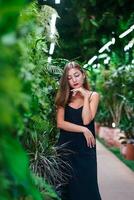 pensando lindo mulher em pé dentro matagal do tropical plantas. jovem mulher vestindo Preto vestir em fino correias carrinhos entre verde plantas dentro estufa. menina com dela olhos baixa pensamento. foto