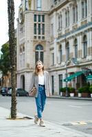 smaling mulher lentamente chacoalhar em rua dentro ensolarado verão dia. jovem mulher indo. menina vestindo Jaqueta e jeans com bolsa. dentro frente do arquitetônico edifícios. velho lindo batumi cidade dentro georgia. foto