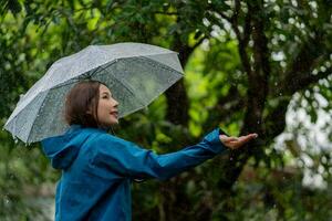 atraente jovem ásia mulher sorrindo carregando guarda-chuva e vestindo dela casaco enquanto caminhando dentro a chuva. foto