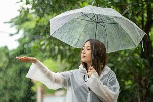 feliz ásia lindo mulher segurando guarda-chuva dentro chovendo estação enquanto em pé dentro a parque. ela tocou a pingos de chuva com dela mão. foto