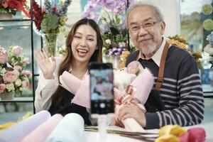 comércio eletrônico negócios, ásia mais velho homem e filha florista mostra floral arranjos e fala para Câmera através da conectados transmissão ao vivo Webcam inscrição dentro uma brilhante flor comprar, lindo flores loja. foto