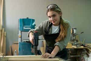 trabalha duro carpinteiro mulher usando Ferramentas sorridente com confiança jovem fêmea marceneiro dentro avental em pé perto bancada e olhando às Câmera amigáveis enquanto trabalhando dentro a construir oficina foto