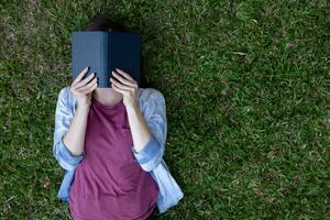 jovem mulher deitado sozinho lendo livro em verde gramado dentro quintal e tendo cópia de espaço para colocar texto em lado em verde gramado a conceito do lendo livros para aprender e Compreendo a artigos dentro a livro. foto