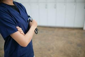 fêmea médico dentro Sombrio azul uniforme carrinhos com dela braços cruzado em borrado fundo do armário depois de examinando paciente dentro emergência sala. fêmea médico dentro uniforme e estetoscópio em borrão fundo foto