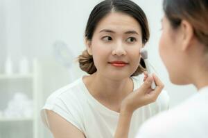 lindo ásia mulher sentar dentro frente do uma espelho e sorrir em inventar. face do uma saudável mulher aplicando inventar. anúncio, estilo de vida , cosméticos, Maquiagem acessórios, beleza atividade, esteticista foto