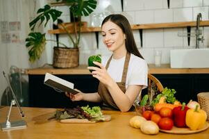 jovem lindo mulher dentro a cozinha dentro a avental, fresco legumes em a mesa, escreve baixa dela favorito receitas, vem acima com Ideias para pratos foto