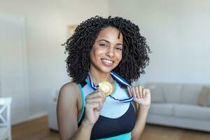 retrato do a atraente jovem fêmea atleta posando com dela ouro medalha. africano americano atleta mostrando primeiro Lugar, colocar medalha foto