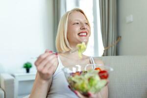 retrato de uma menina lúdica feliz comendo salada fresca de uma tigela na cozinha dela. bela mulher em forma comendo salada saudável após treino de fitness foto