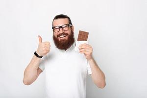 retrato de um homem barbudo alegre em uma camiseta branca mostrando o polegar para cima e uma barra de chocolate