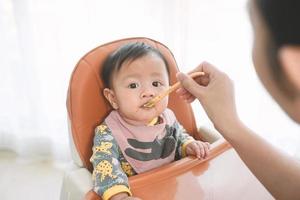 Menina de 6 meses de idade comendo mistura de comida em uma cadeira alta. foto