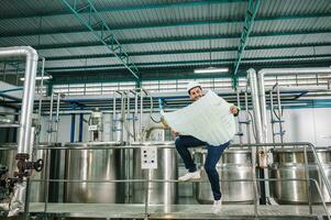 caucasiano técnico engenheiro homem sentado e segurando projeto do industrial projeto com caldeira e gasoduto dentro bebida em processamento plantar foto