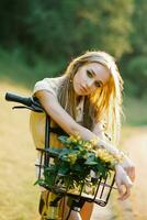 uma jovem lindo mulher detém uma bicicleta com amarelo verbena flores dentro uma cesta em a ao ar livre país andar foto