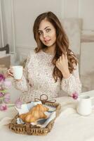 uma cabelos castanhos mulher com grandes cabelo é tendo café da manhã às a mesa com croissants e uma copo do café. Primavera retrato. foto