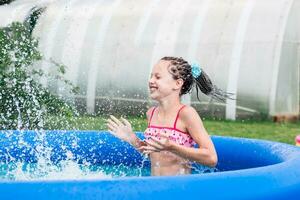 alegre pequeno menina com tranças afro salpicos água dentro a inflável piscina em uma verão dia dentro a quintal foto