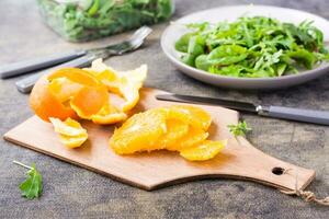 descascado e fatiado laranja em uma corte borda e uma misturar do Rúcula, acelga e mizun em uma prato em a mesa. ingredientes para fazer Vitamina dietético vegetariano salada foto