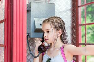 uma sério menina com tranças afro faz uma telefone ligar dentro a velho Inglês Telefone cabine. geracional contraste foto